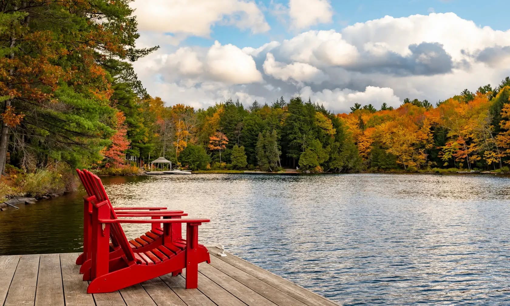 Red Muskoka chairs on a dock overlooking a lake framed by autumn trees